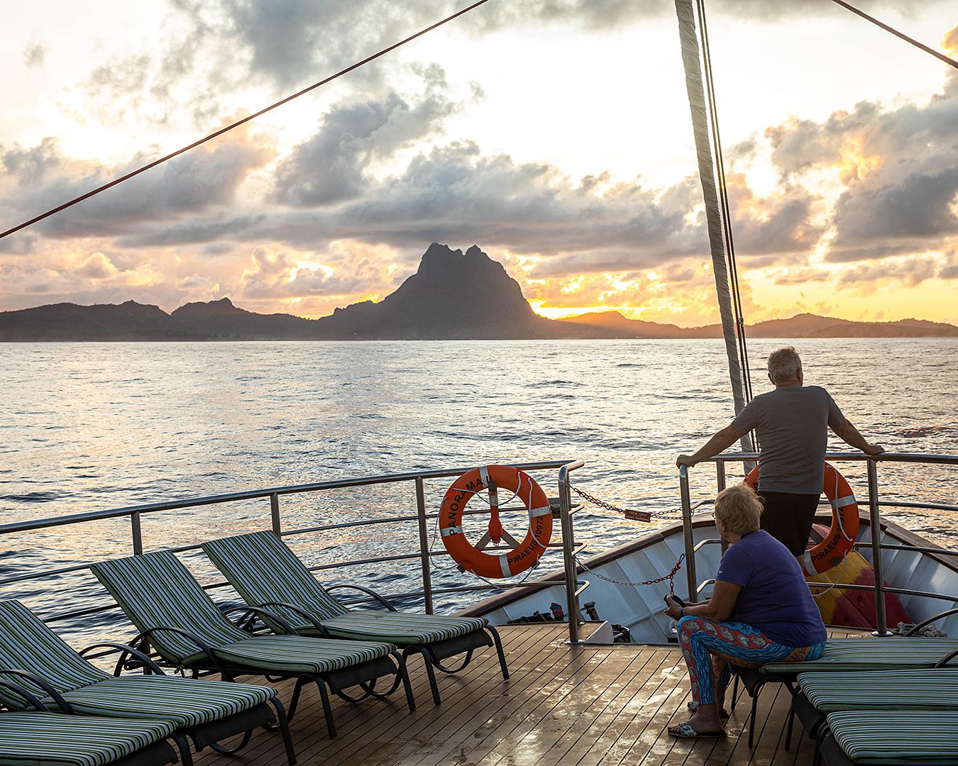 Passageiros relaxando nas espreguiçadeiras do deck de um navio observando o pôr do sol atrás de montanhas no horizonte marinho.