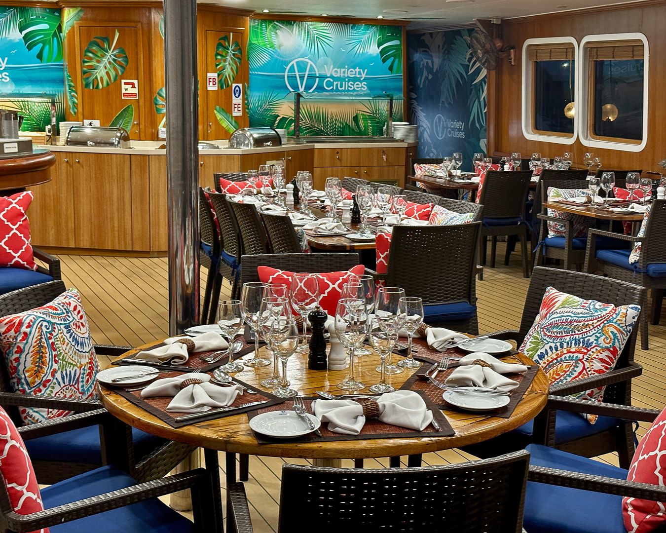 Salão de jantar refinado a bordo de um navio com mesas postas, taças de vidro, cadeiras confortáveis e decoração tropical.