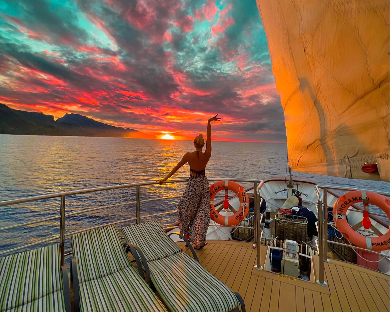 Mulher de costas no deck de um veleiro contemplando um pôr do sol colorido em tons de laranja e rosa no oceano.