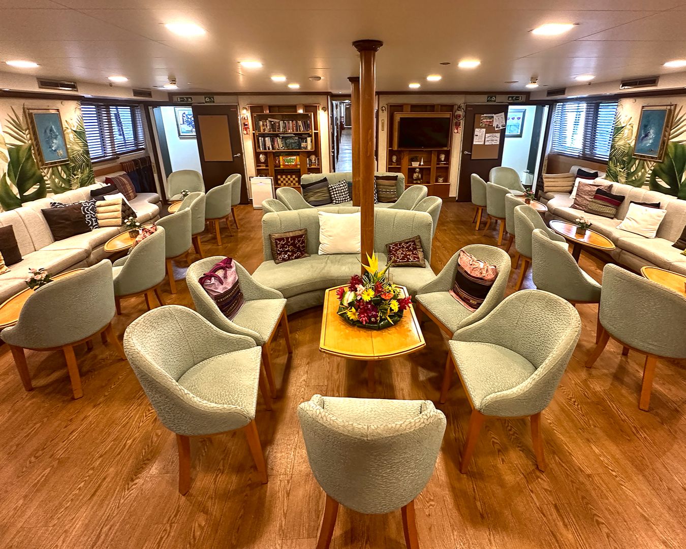 Lounge interno de um cruzeiro de luxo com poltronas verdes, sofás confortáveis, piso de madeira e arranjo de flores tropicais ao centro.