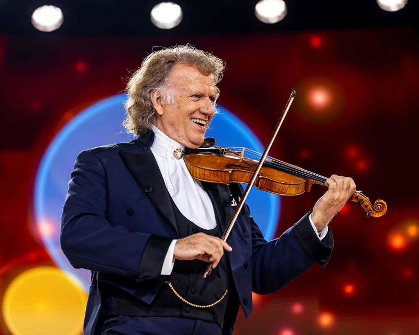 O maestro e violinista André Rieu sorrindo enquanto toca seu violino durante um show ao vivo