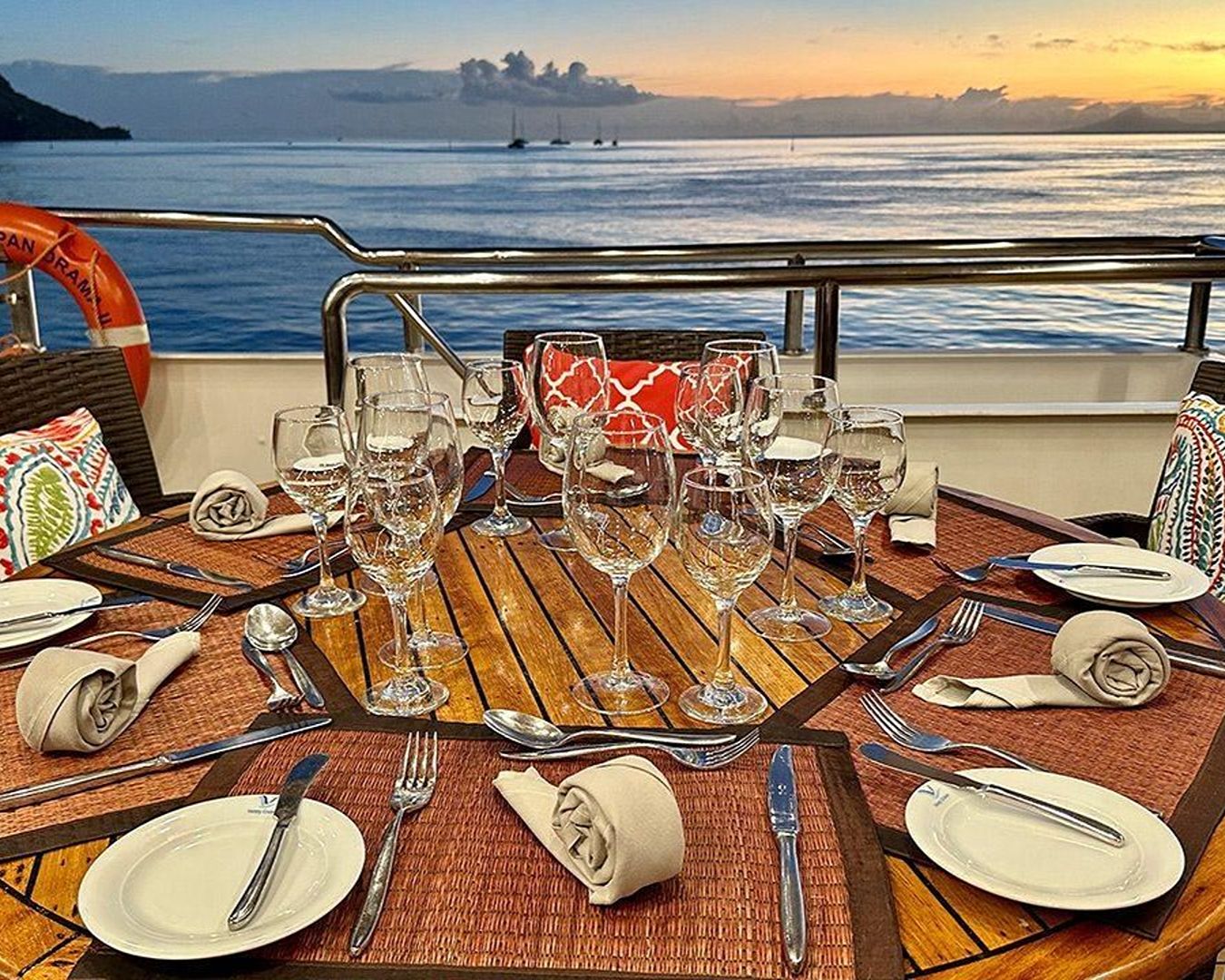 Mesa redonda de madeira posta no deck externo de um navio com taças de vinho, talheres e guardanapos diante de um pôr do sol no mar.