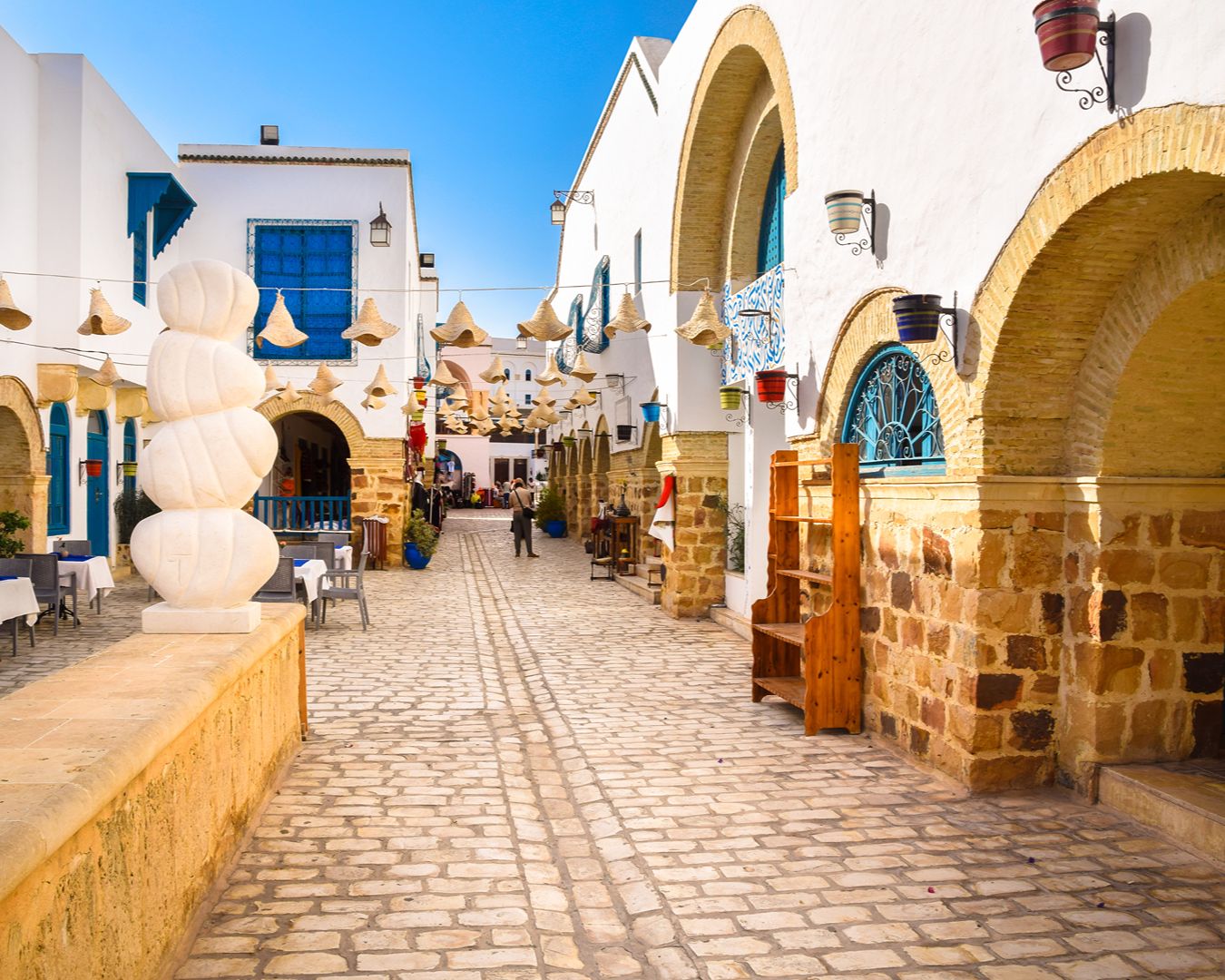 Rua de vilarejo tradicional com arcos de pedra, paredes brancas, esculturas e luminárias artesanais na Tunísia