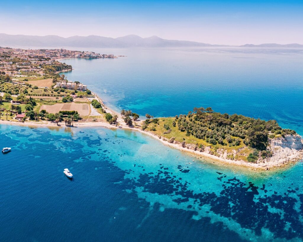 Vista aérea de península com praia de águas cristalinas, barcos ancorados e vegetação costeira na Grécia