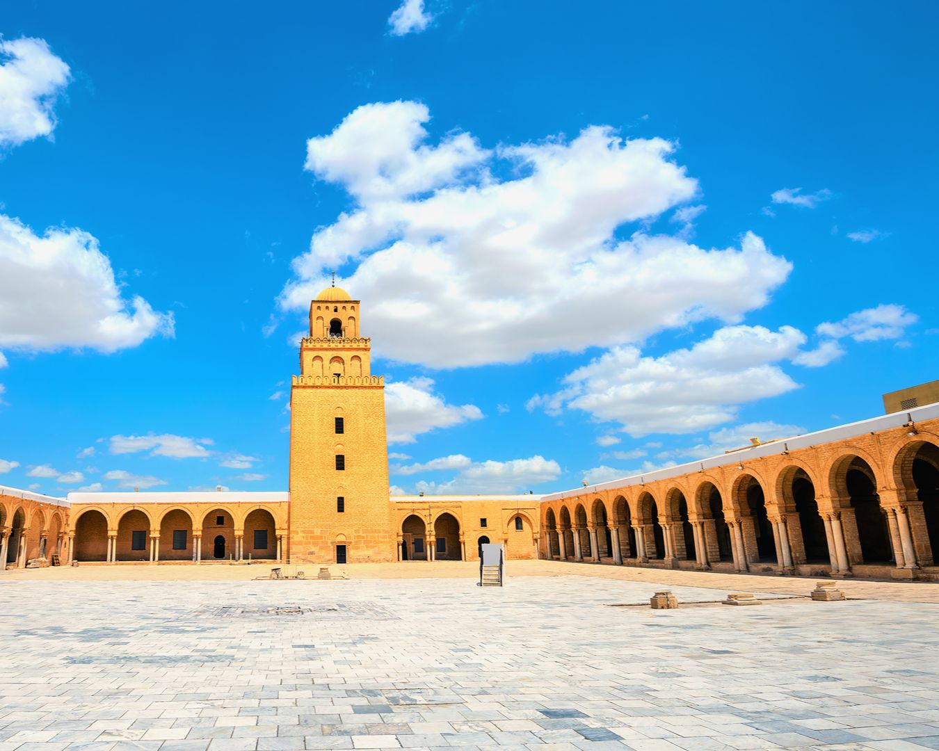 Pátio interno de mármore e minarete da Grande Mesquita de Kairouan sob céu azul com nuvens