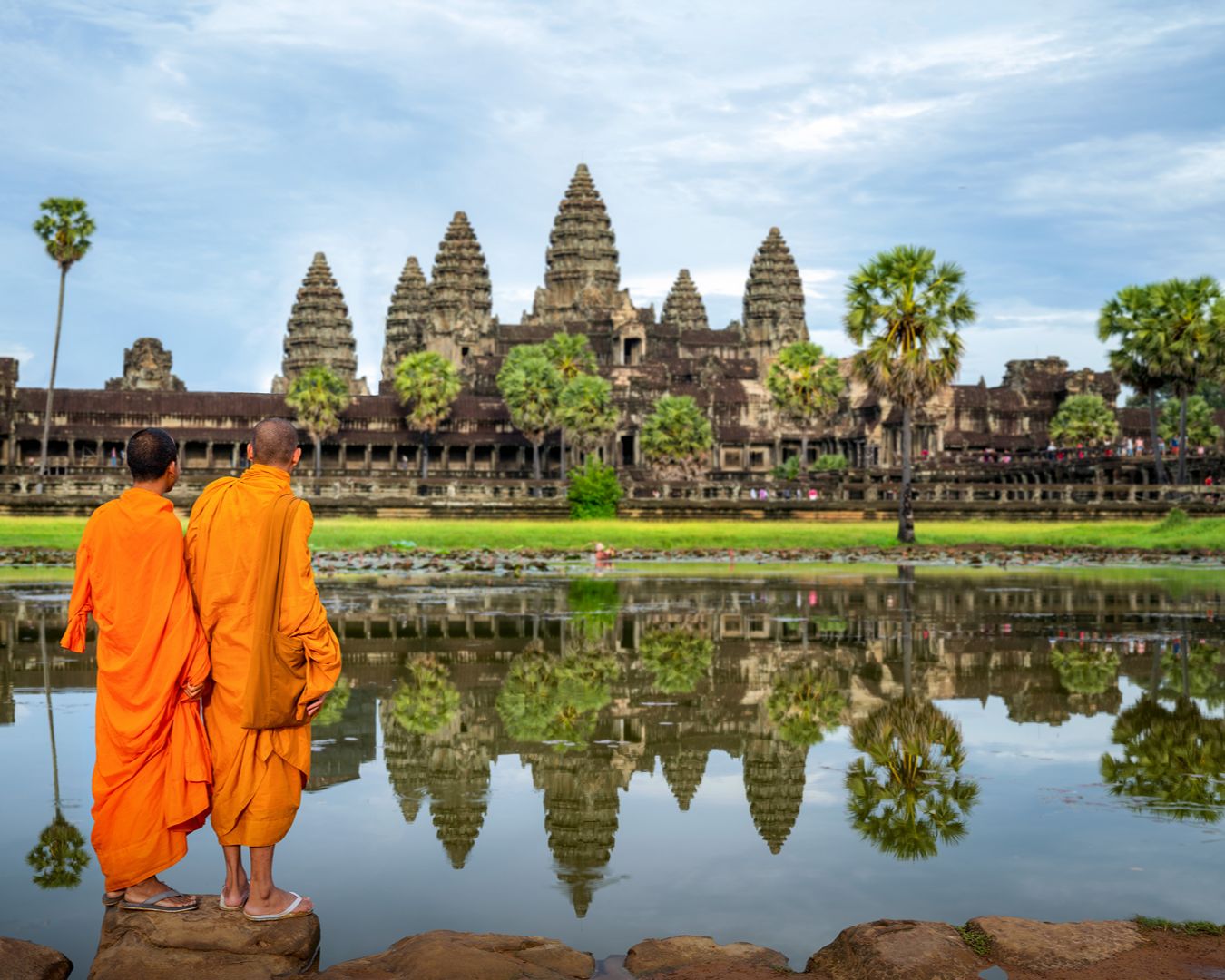 Dois monges budistas com trajes laranjas observando o reflexo do templo Angkor Wat no lago