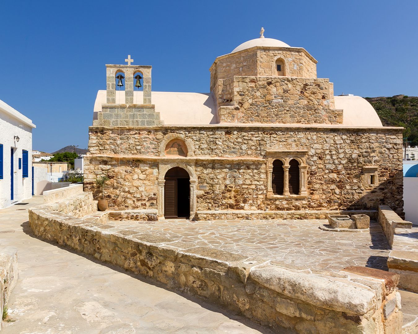 Fachada de uma antiga igreja de pedra com campanário e cúpula em uma vila grega sob céu limpo