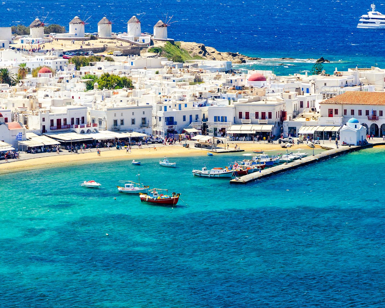 Vista aérea do porto de Mykonos com águas cristalinas, barcos ancorados e os famosos moinhos de vento ao fundo